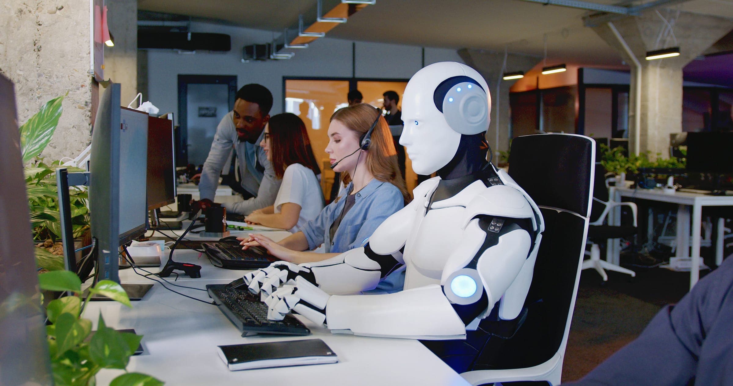 A humanoid robot typing at a desk beside people in an open office