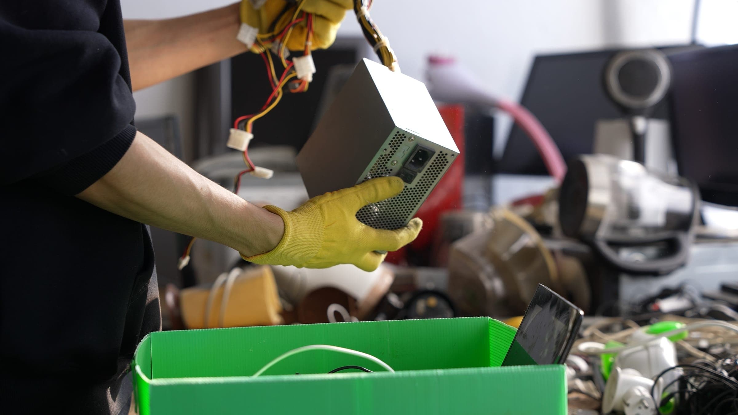 Gloved hands sorting a computer power supply into a recycling bin of old electronics